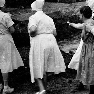 Here's a brief summary of the image: Women in uniforms observe an excavation site outdoors.