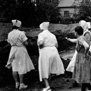 Here's a brief summary of the image: Women in uniforms observe an excavation site outdoors.