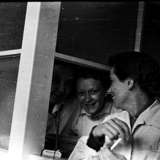 Two women laughing indoors near open window.