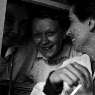 Two women laughing indoors near open window.