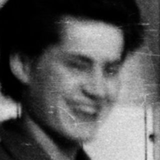 Person smiling while holding a suitcase in hallway.