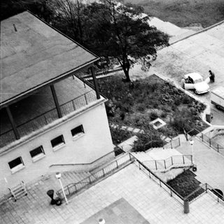 Aerial view of mid-century building with stairs, parked vintage vehicles, and people.