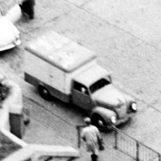 Aerial view of mid-century building with stairs, parked vintage vehicles, and people.