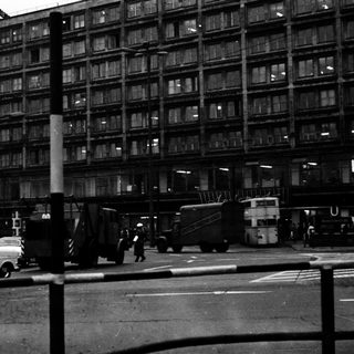 Urban street scene with mid-century buildings, vintage cars, and pedestrians.