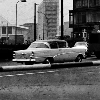 Urban street scene with mid-century buildings, vintage cars, and pedestrians.