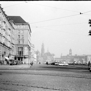 Mid-20th-century city street with vintage cars, buildings, and cathedral in background.