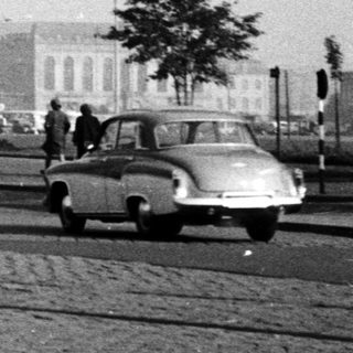 Mid-20th-century city street with vintage cars, buildings, and cathedral in background.