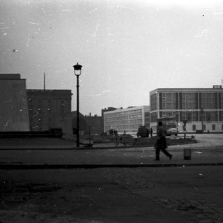 Urban street scene with mid-century buildings, lone pedestrian, and horse-drawn carriage.