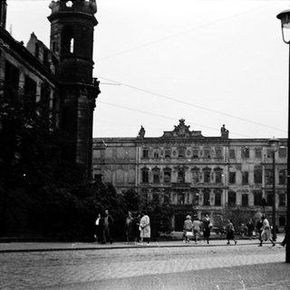 Historic city square with grand damaged building and pedestrians.
