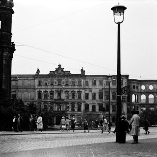 Historic city square with grand damaged building and pedestrians.