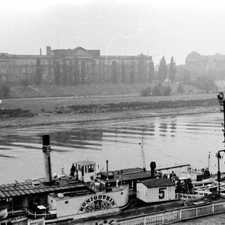 Steam-powered barge on river near historic buildings.