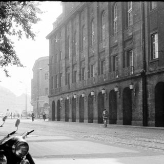 Mid-century urban street with parked motorcycles, vintage cars, and large institutional building.