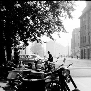 Mid-century urban street with parked motorcycles, vintage cars, and large institutional building.