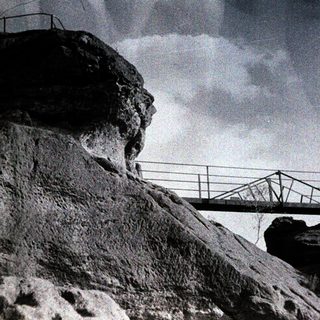 Dramatic bridge spanning rocky cliff under stormy sky.