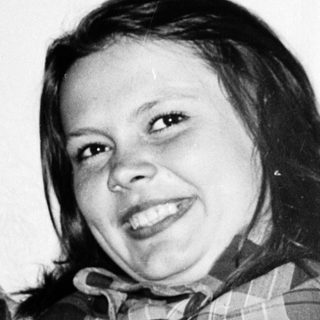 Smiling person wearing plaid shirt, seated indoors.