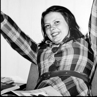Smiling person wearing plaid shirt, seated indoors.