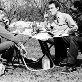 Three men camping, eating outdoors by a fire.