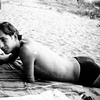 Shirtless man resting on towel near water.