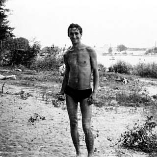 Shirtless man standing by waterfront in shorts.