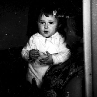 Young child in white outfit holding object near window.