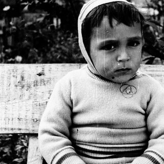Young child sitting outdoors, wearing sweater and hat.