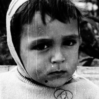 Young child sitting outdoors, wearing sweater and hat.