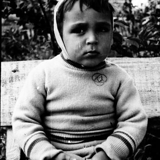 Young child sitting outdoors, wearing sweater and head covering.