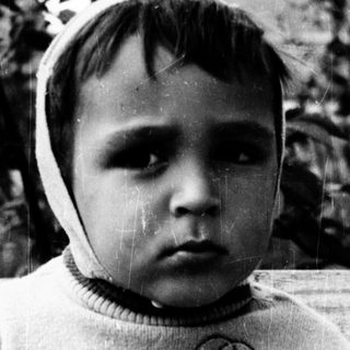 Young child sitting outdoors, wearing sweater and head covering.