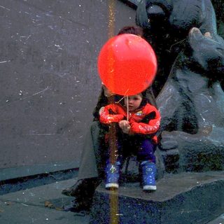 Child blowing red balloon near bear statue.