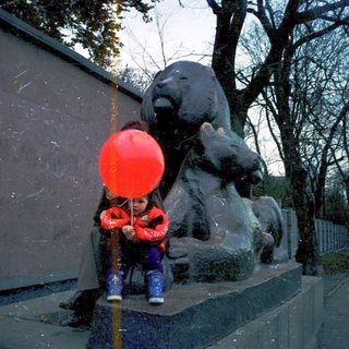 Child blowing red balloon near bear statue.