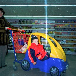 A woman pushes a shopping cart while a child rides a toy car inside.