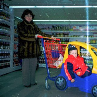 A woman pushes a shopping cart while a child rides a toy car inside.