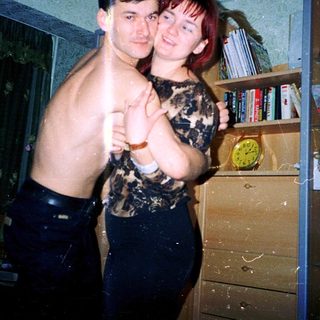 Two people posing indoors, shirtless and holding hands.