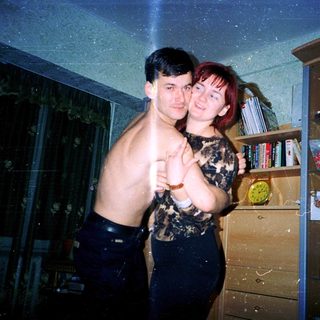 Two people posing indoors, shirtless and holding hands.
