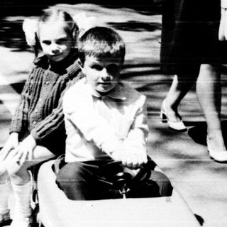 Two children riding toy cars outdoors.