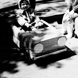 Two children riding toy cars outdoors.
