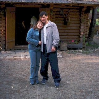 Two people posing outside rustic wooden cabin in forest.
