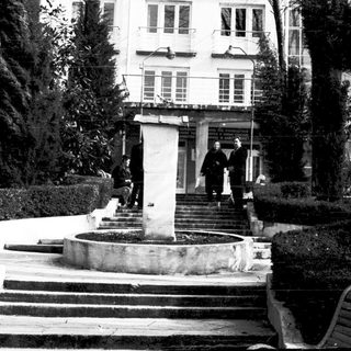 Black-and-white photo of people near a building entrance with steps and fountain.