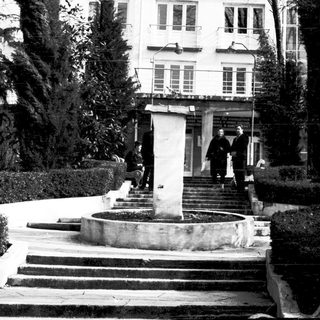 Black-and-white photo of people near a building entrance with steps and fountain.