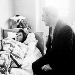 Man reading newspaper to sick woman in bed.