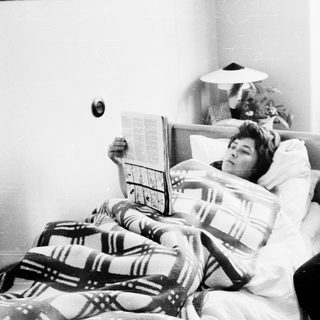 Man reading newspaper to sick woman in bed.