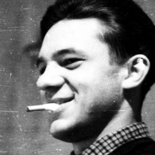 Young man smoking cigarette indoors against plain wall.