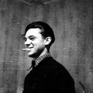 Young man smoking cigarette indoors against plain wall.