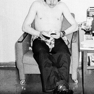 Shirtless man shaving in a cluttered room.