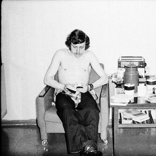 Shirtless man shaving in a cluttered room.