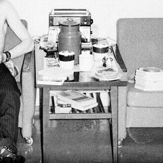 Shirtless man shaving in a cluttered room.