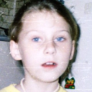 Child holding toy near refrigerator in cluttered room.