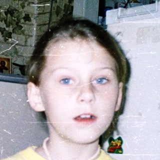 Child holding toy near refrigerator in cluttered room.