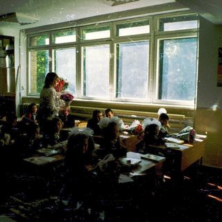Classroom with students writing at desks, teacher standing.