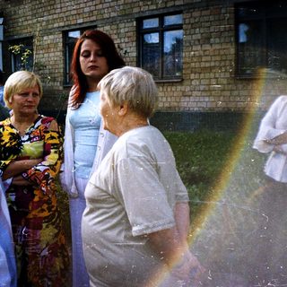 People gathered outdoors near a building during rain with rainbow.